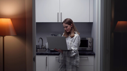 Fototapeta na wymiar Young woman in home wear using laptop standing in kitchen