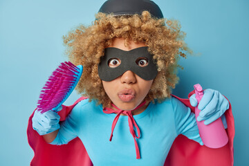 Studio shot of amazed woman with curly hair pretends being superhero holds brush and bottle of detergent has wondered expression does housework isolated over blue background. Cleanliness concept