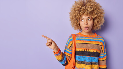 Stunned shocked female model with curly hair gasps impressed says wow points index finger on blank space shows something breathtaking isolated over purple background. Look at sale logo or banner