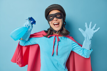 Joyful Asian woman dressed in superhero costume holds spray detergent looks gladfully above keeps hand raised ready for cleaning house isolated over blue background. Household chores concept