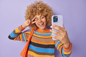 Beautiful positive woman makes peace gesture over eye sticks out tongue winks eye poses at smartphone camera makes selfie dressed in knitted jumper carries small bag isolated over purple wall