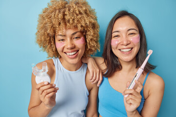Happy mixed race young women hold mouth expander and electric toothbrush smiles positively apply hydrogel patches under eyes to remove puffiness undergo daily hygiene and beauty routines at home