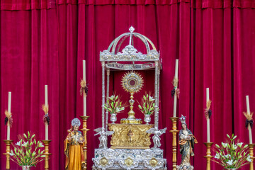 Image of Corpus Christi in gold and silver between candles and flowers with a red curtain in the background