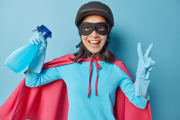 Positive superwoman makes peace gesture holds bottle of detergent ready for cleaning house wears protective helmet mask cloak and rubber gloves isolated over blue background. Feminine power.