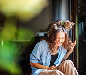 Beautiful young woman laughing happily with a cat on her head, companion pet friendship concept
