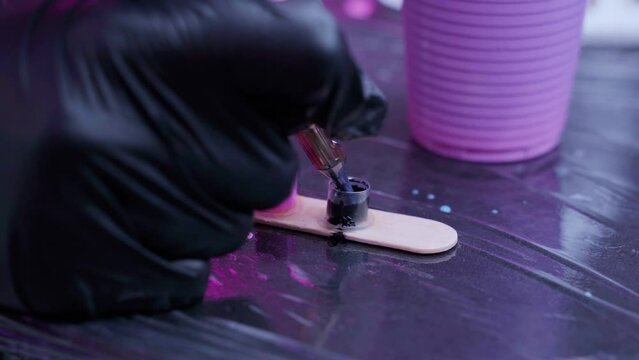 Close-up hand tattoo artist in black medical glove dips tattoo machine needle in black paint against the background of special tattoo tools in pink light