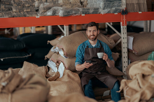 Barista sitting on the warehouse with bags of coffee beans, drinking coffee and using phone