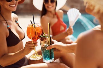 Close up of female friends toasting while drinking cocktails at swimming pool during summer day.