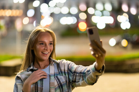 Closeup cheerful girl taking selfie photo by mobile phone on night city street. Portrait of happy woman showing victory sign to smartphone camera outdoor. Smiling lady looking cellphone camera outside