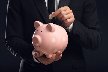 Businessman putting coin in piggy bank on black background, closeup