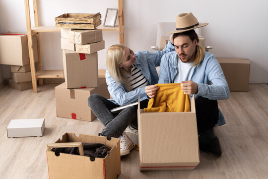 Young couple unpacking boxes with things and clothes moving to a new house, new apartment owners sitting on the floor with boxes, happy moving time