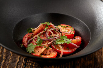 Traditional uzbek salad with tomatoes and sweet onion on black bowl. Tomatoes and onion salad on wooden background in rustic style. Appetizer from tomato and coriander.