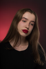 Portrait of a young girl with a dreamy look in a black blouse on a red background. Long brown hair lies loosely on the shoulders. Expressive, plump lips are painted with red lipstick. Close-up.