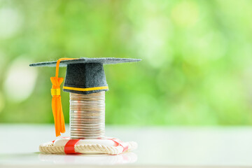 Tuition protection service and tuition refund insurance, financial concept : Black graduation cap or a mortarboard placed higher on top of a coin stack with a red lifebuoy on a table, green background - obrazy, fototapety, plakaty