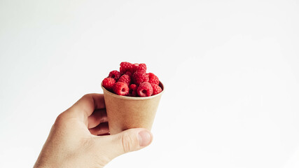 Fresh raspberries in a paper cup held by a male hand on a white background