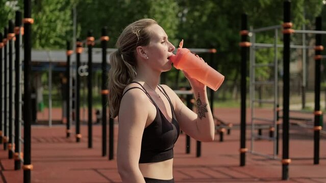 Young woman drinking water from bottle at sports ground after training. Rest after hard workout. Relax time. Female taking break after workout. 4K, UHD