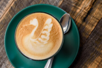 A latte foam art on a cup of coffee, placed in a drinking glass and green saucer with a wood grain background
