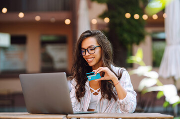 Young woman sitting at cafe making online shopping, using credit card and laptop. Black friday, sale, consumerist, lifestyle concept. Online shopping. - obrazy, fototapety, plakaty