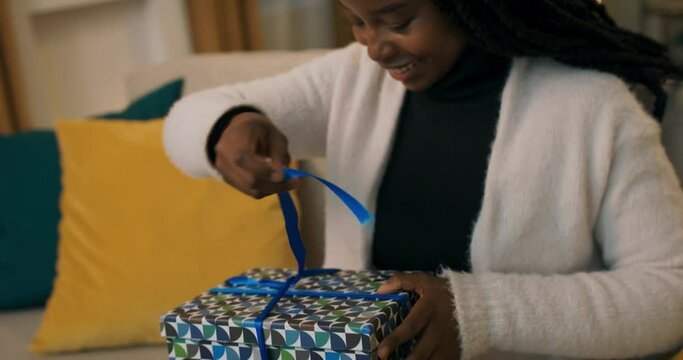 Close-up of an African-looking girl unpacking gift. The girl unties the blue ribbon, opens the box and is infinitely happy with the gift. She clapped hands in joy and smiled.