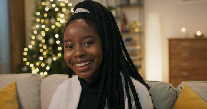 Close-up of young girl of African appearance. The beauty is sitting on the couch and dressed in black golf. She looks at the camera and smiles sincerely. A Christmas tree flickers behind.