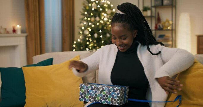 An African-looking girl with pigtails sits on sofa in the living room and holds gift. She unties the blue ribbon and opens the box. The girl was disappointed, because she received unwanted gift.