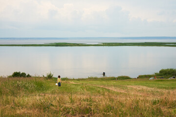A picturesque bank of a wide river with rare vacationers. Clouds are reflected transparently in the water. On the shore in the grass, a child collects a wild berry.