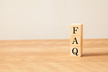 FAQの文字。よくある質問。Frequently Asked Questions。3つの木製ブロックに書かれている。黒い文字。木製テーブルと白い壁紙の背景。左にコピースペース。