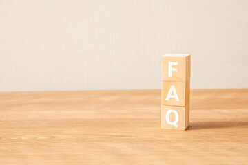 FAQの文字。よくある質問。Frequently Asked Questions。3つの木製ブロックに書かれている。白い文字。木製テーブルと白い壁紙の背景。左にコピースペース。