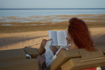 Beauttiful young woman relax at the beach and reading a book. Red hair woman reading book relaxed in deck chair. Summer holiday vacation.                               