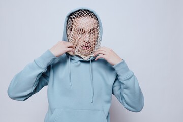 a man with a mesh on his face stands in a light blue hoodie on a light background and tries to...