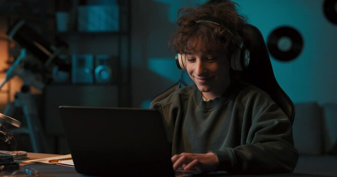 A young boy with curly hair is happy with remote learning. A student sits in front of a computer with wireless headphones listening to a teacher lecture. The lessons take place over the Internet.