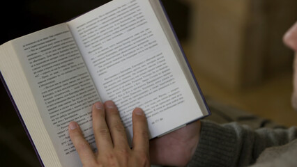 Naklejka premium Over the shoulder view of a man reading a book at home. Video. Middle aged male sitting and reading a paper book, turning pages, relaxing at home.