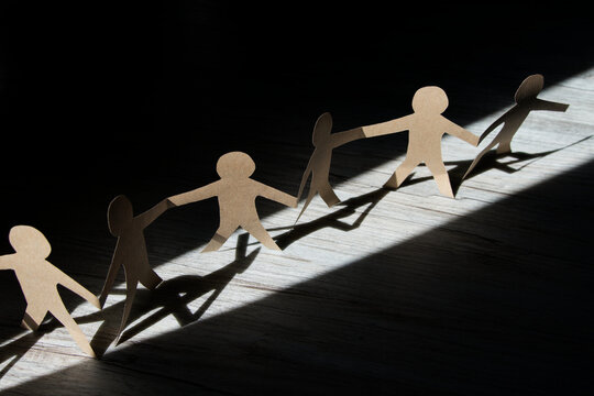 Scherenschnitt Papiermenschenkette aus brauner Pappe im Lichtkegel, schwarzer Hintergrund, horizontal