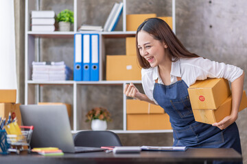 Startup SME small business entrepreneur of freelance Asian woman using a laptop with box Cheerful success happy Asian woman her hand lifts up online marketing packaging and delivery SME idea concept