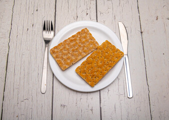 two slices of crispbread on a plain white plate and cutlery with signs of use on a rustic wooden surface