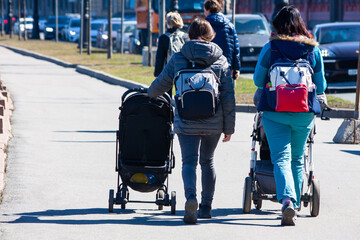 Two mothers with prams and backpacks walk with children around the city