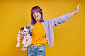 Happy young woman carrying little dog and gesturing while standing against yellow background