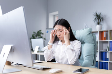 Exhaustion at work. A young Asian woman holds her head, feels pain, grimaces. Sitting in the office at the desk, working at the computer, exhausted. Businesswoman, accountant, freelancer.