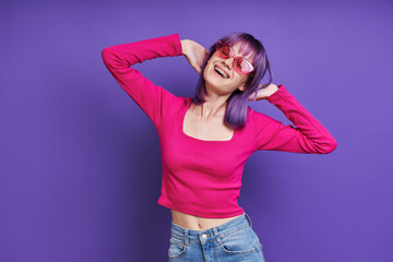 Cheerful young woman holding hands behind head while standing against purple background