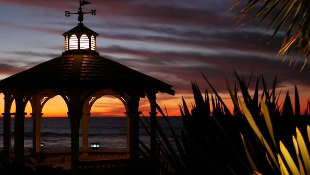 Sunset cloudscape and gazebo silhouette on beach, twilight dusk, clouds in dramatic purple pink sky, California coast. Wooden romantic cosy alcove and palm tree. Weather wind vane or roof weathercock.