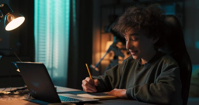 Teenage boy doing homework using computer sitting by desk in room alone. Teenager watching video and browsing internet on laptop, webcam talking.