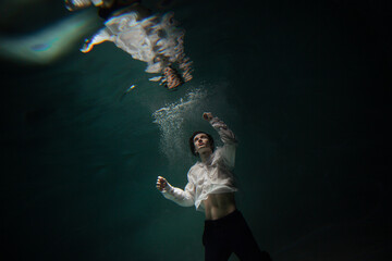 Naklejka na ściany i meble underwater shooting with contrasting light, a guy in a white shirt and pants screaming underwater, panicking and afraid of drowning, falling into the water, a crime.