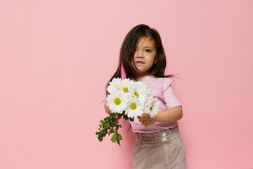 a cute beautiful little preschool girl stands on a pink background in a pink T-shirt and holds a small bouquet of white daisies in her hands, smiling cheerfully and holding it out a little forward