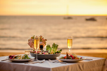 Romantic sunset dinner on beach. Table honeymoon set for two with luxurious food, glasses of champagne drinks in restaurant with sea view and yachts on background. Summer vacation or wedding concept.