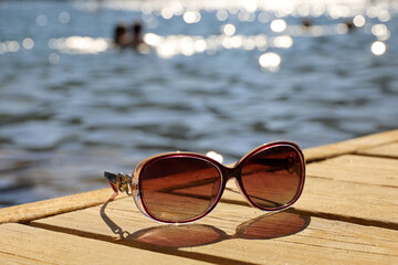 Fototapeta na wymiar Female sunglasses on wooden boards on sea and swimming people background. Beach vacation in summer, defocused view to shining water