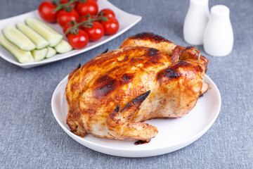 Chicken baked whole to a delicious crust. A traditional dish. In the background are cucumbers and cherry tomatoes on a white plate. Close-up, selective focus, gray background.