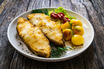 Fish dish - fried halibut with fried potatoes, cherry tomatoes and leafy greens on wooden table
