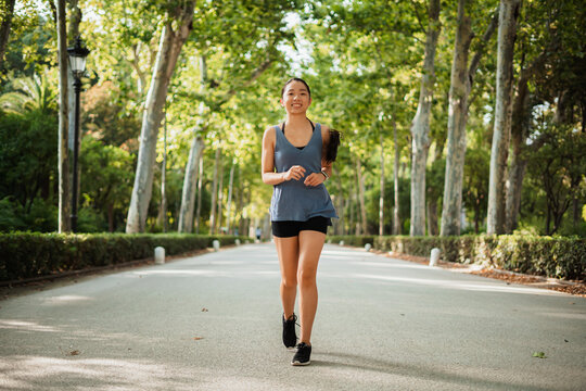 Cheerful fitness young woman running in the park surrounded by green trees