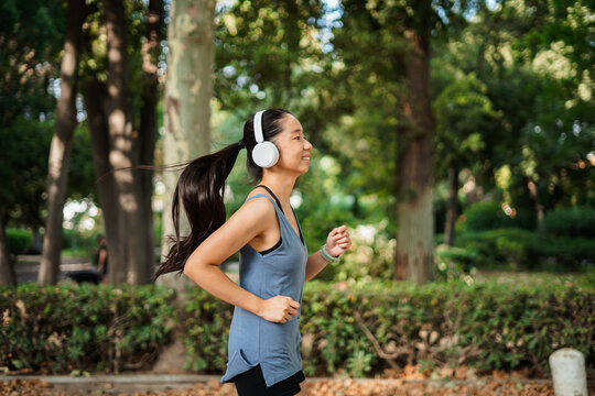 Happy asian athlete girl running in the park while she is listening to music on white headphones