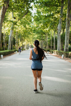 Vertical back shot of athlete girl jogging in the park surrounded by green trees.